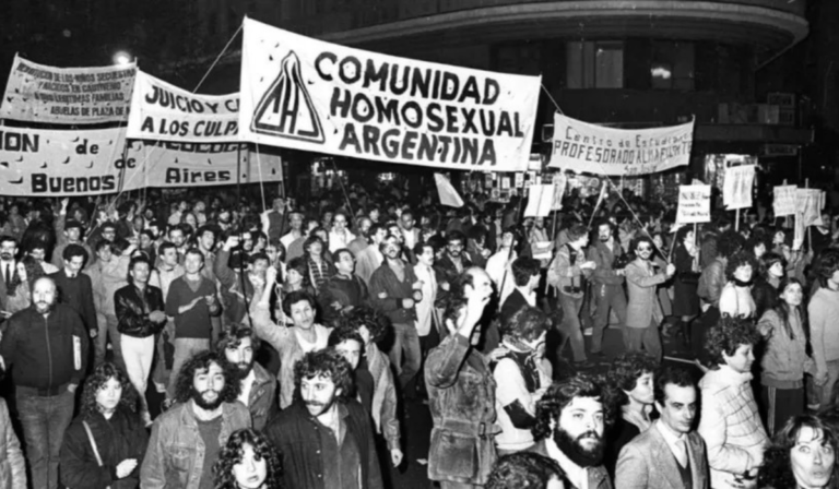 Fotografía de primera marcha LGBT en Argentina. Se ven personas marchando y la bandera de Comunidad Homosexual Argentina