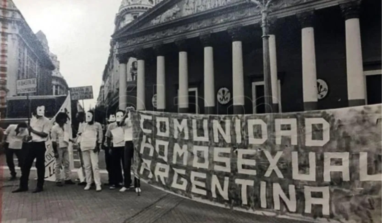 Fotografía de primera marcha LGBT en Argentina. Se ve la bandera de Comunidad Homosexual Argentina y activistas con máscaras blancas.